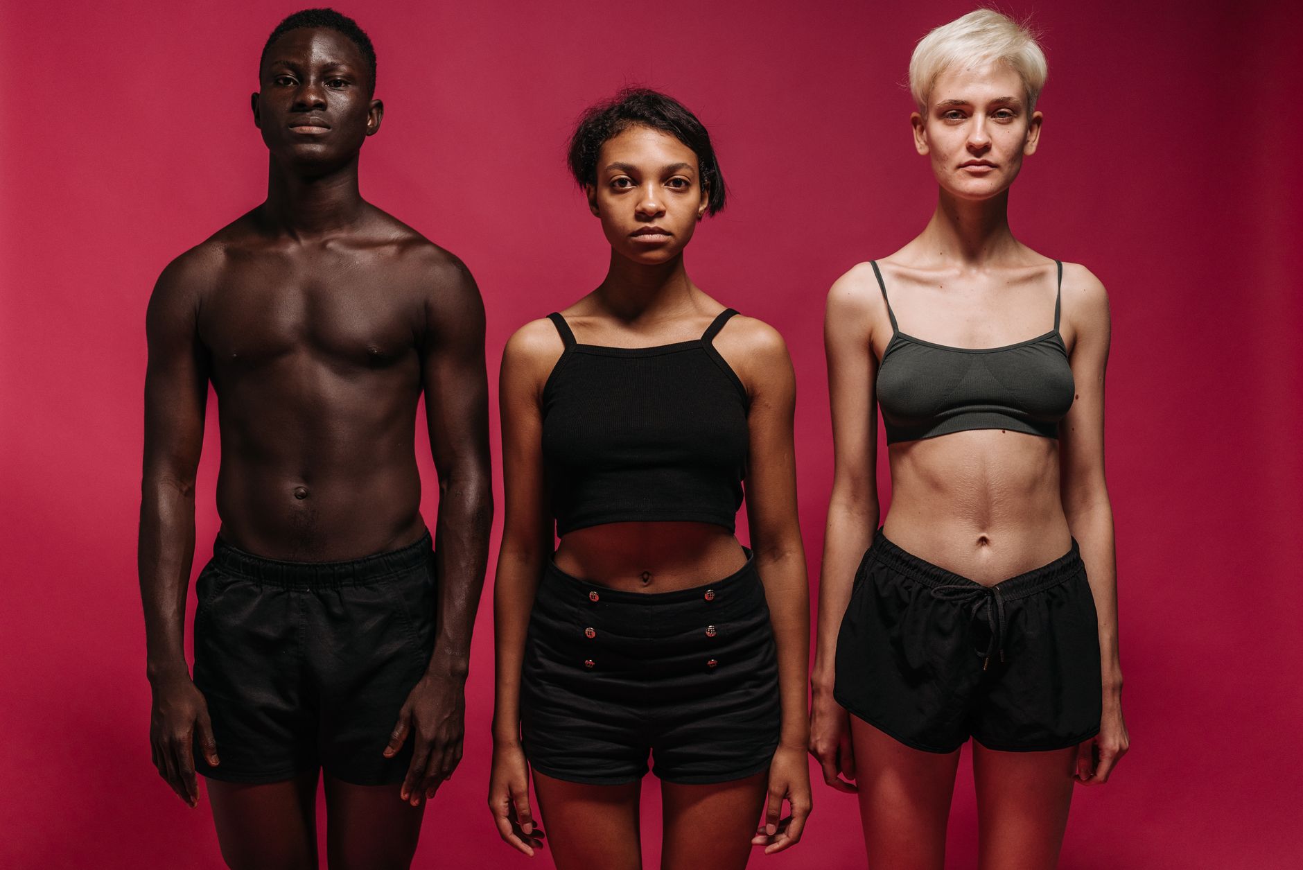 Three diverse individuals stand confidently against a pink background in fitness attire.