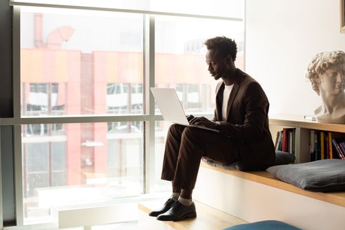 Free Adult black man working on a laptop in a stylish, modern office setting with city views. Stock Photo