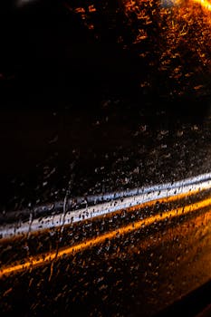 Moody shot of rain streaks and light reflections on a car window during night drive.