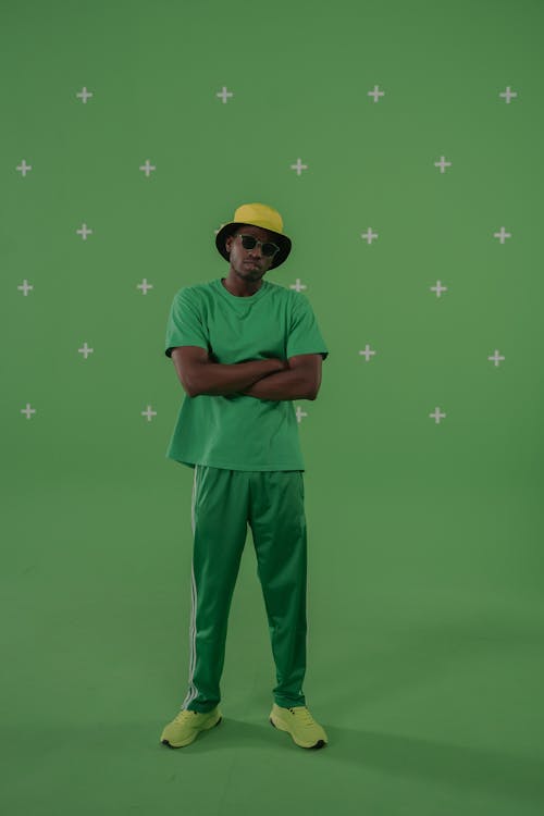 A Man in Orange Bucket Hat Wearing Green Shirt and Black Pants while Standing Near Green Wall