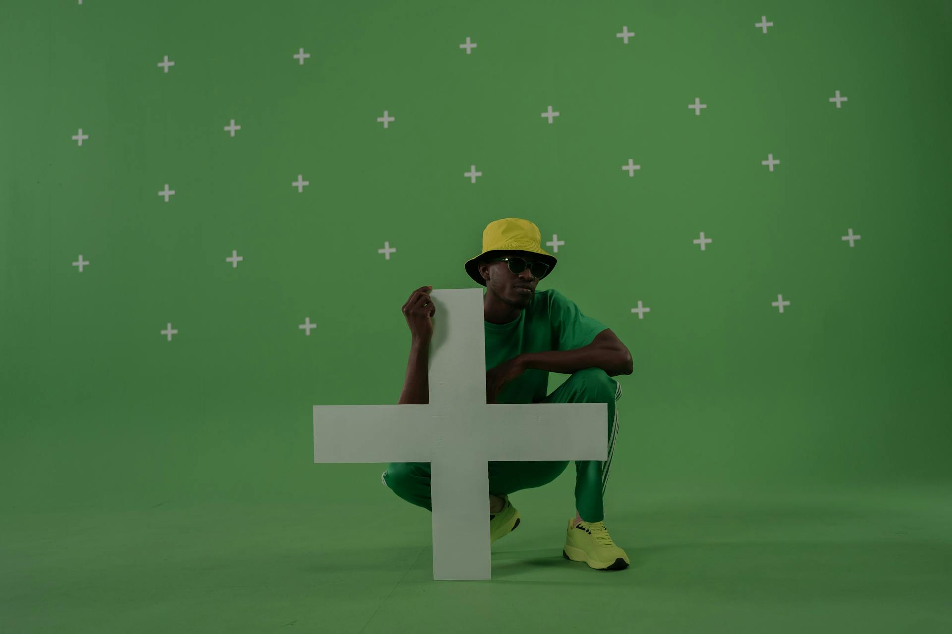 Crouching man in a studio with green chroma key and plus symbol.