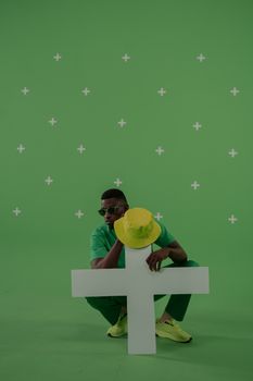 Stylish man poses with a yellow bucket hat and cross. Green background adds a vibrant touch.