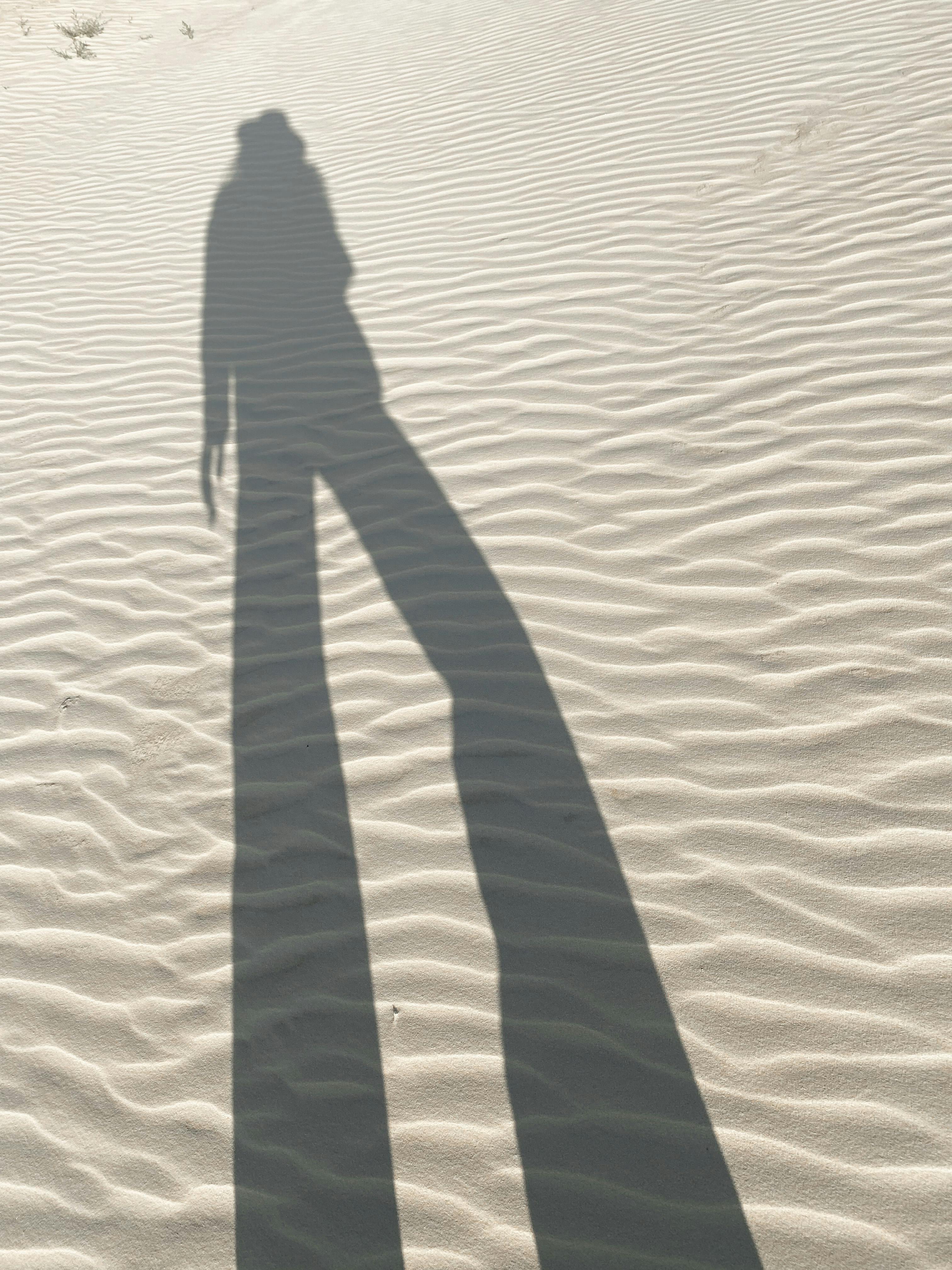 Tall shadow of a person cast on rippled sand dunes under sunlight.