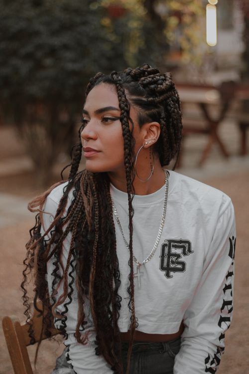Free A fashionable young woman with braided hair sitting outdoors, wearing a crop top and silver jewelry. Stock Photo