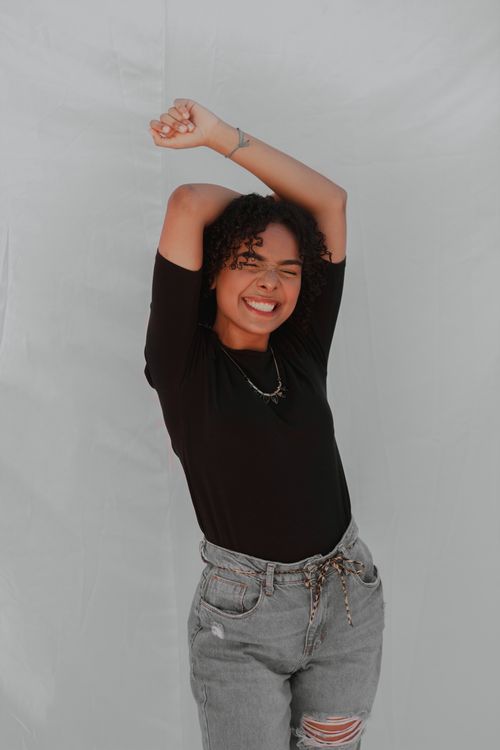 Free Smiling woman poses with arms raised, wearing a black shirt and ripped jeans against a white backdrop. Stock Photo