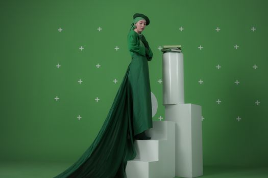 Elegant woman in a green dress standing on steps with books, against a green background.