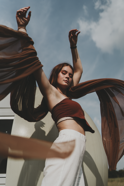 Fashionable woman in brown attire poses with a flowing scarf, set against a sunny outdoor backdrop.