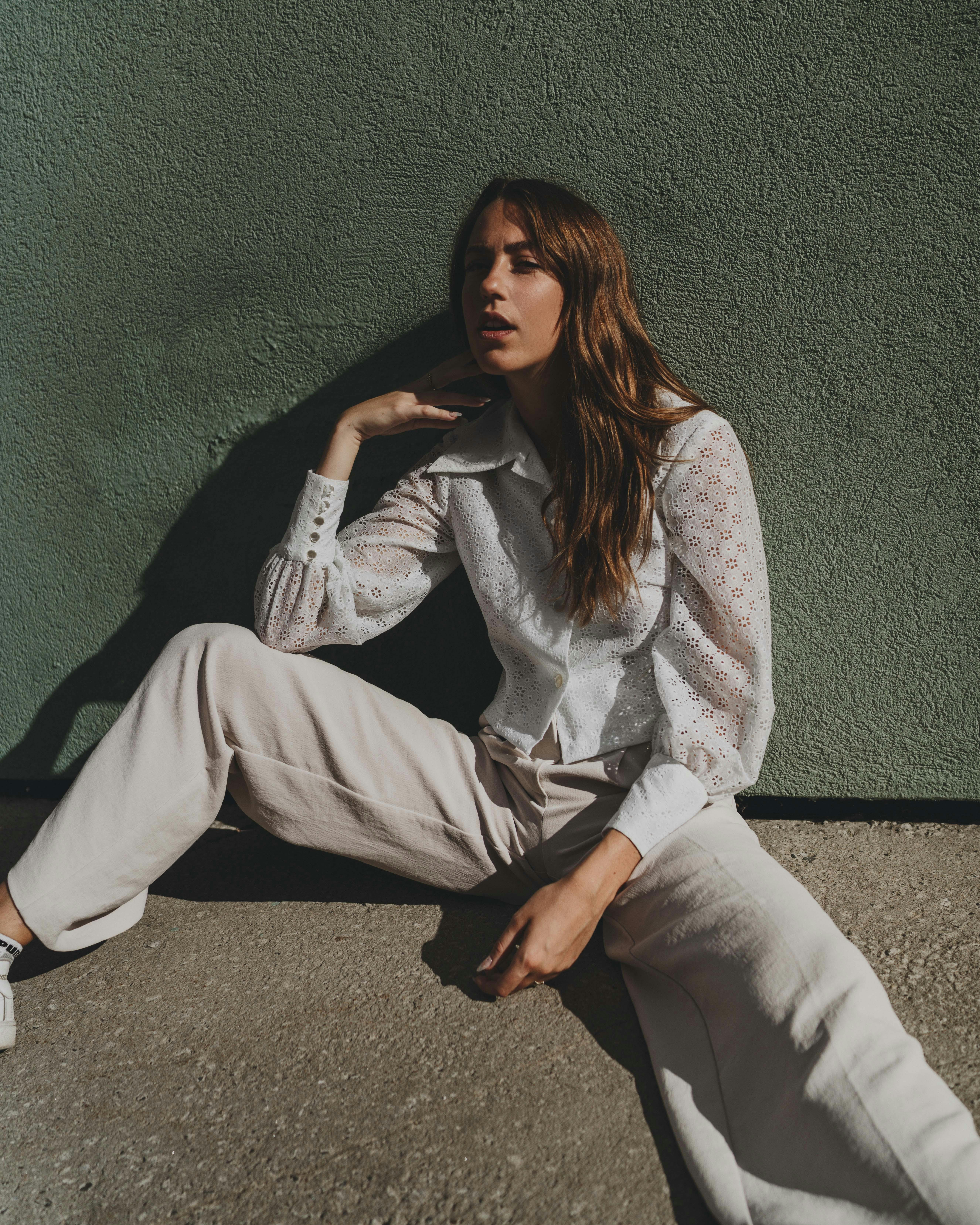 Stylish woman in a white blouse and pants sits elegantly against a textured wall.