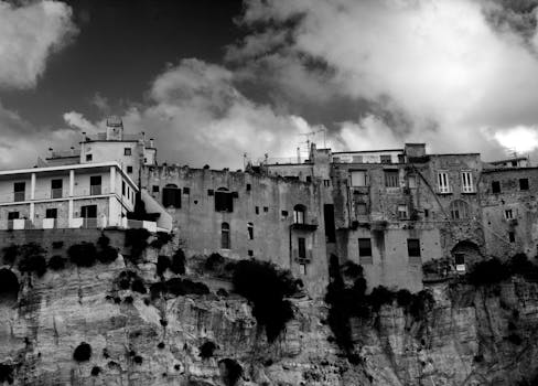 Black and white view of dramatic clifftop buildings in Tropea, Italy against a cloudy sky.