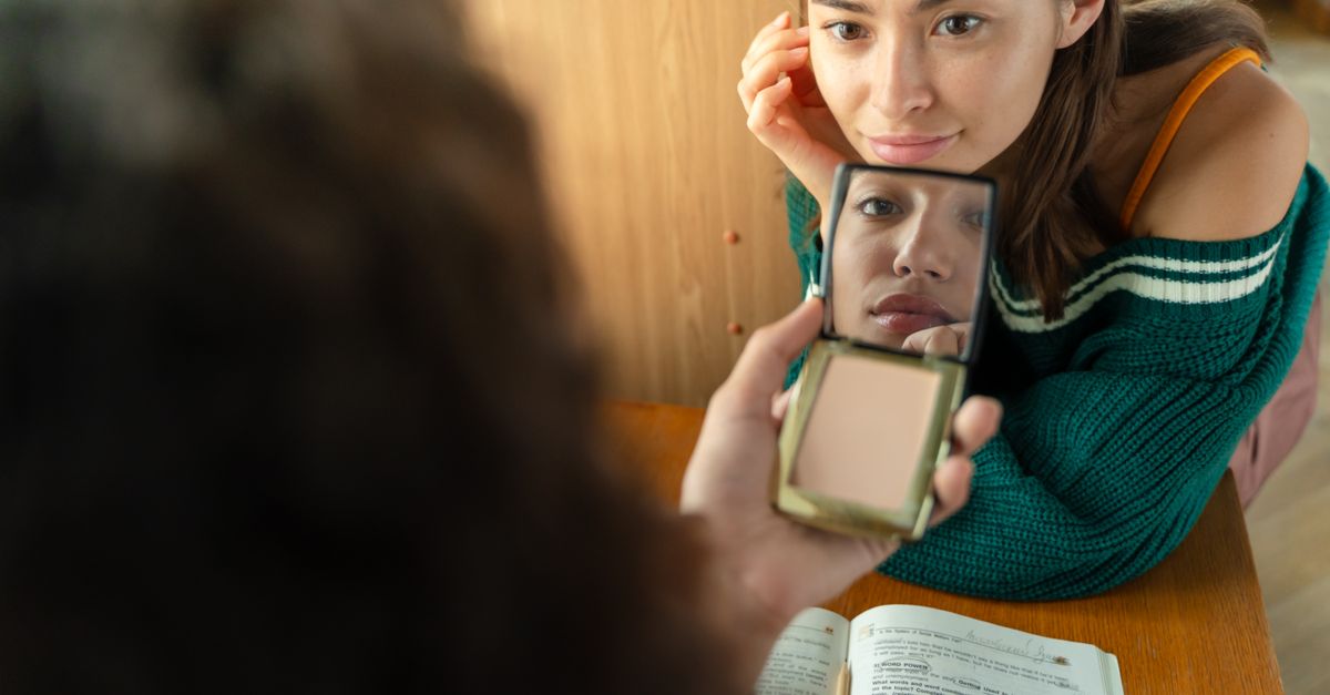 A young woman looks into a mirror while studying at a desk, reflecting thoughtfulness.