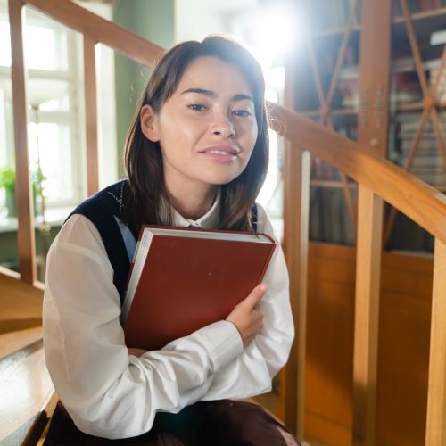 Free A young woman sits on a library staircase holding a book, surrounded by warm interior lighting. Stock Photo
