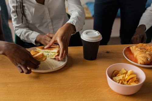 Free Close-up of a diverse group sharing sandwiches, coffee, and snacks at a wooden table. Stock Photo