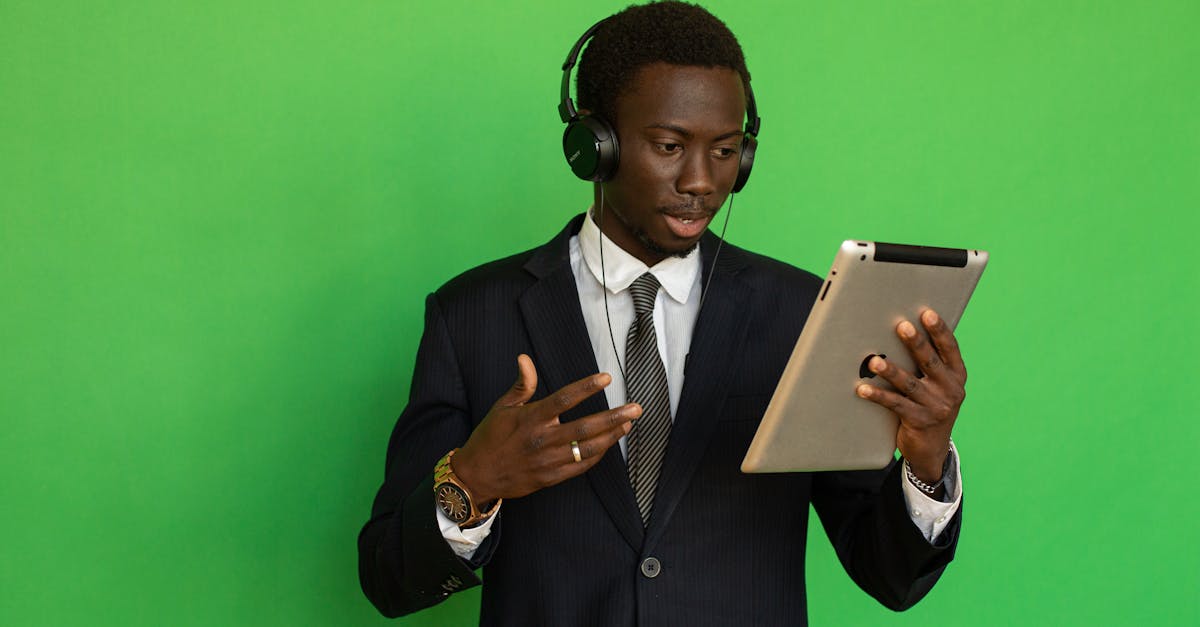 Young black man in formal attire using a tablet with headphones against a green background.