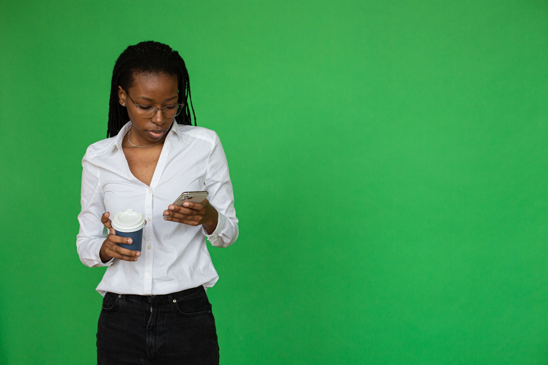 Businesswoman holding coffee and using her phone with a green screen background, ideal for digital projects.