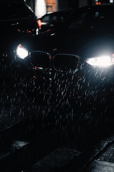 A dark street with bright car headlights shining through the rain at night, creating a moody urban atmosphere.
