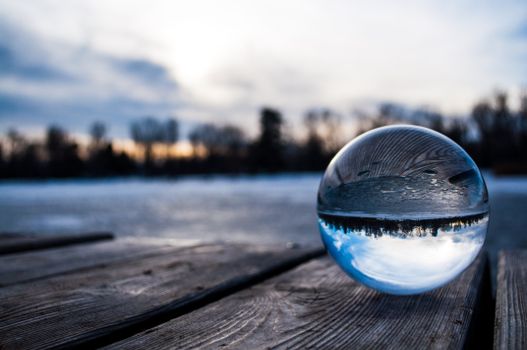 A serene reflection of a winter sunset captured in a glass sphere on a wooden deck by a frozen lake.