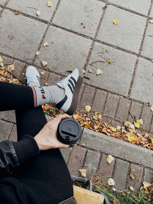Adult wearing sneakers and holding coffee on a fall day, surrounded by fallen leaves.
