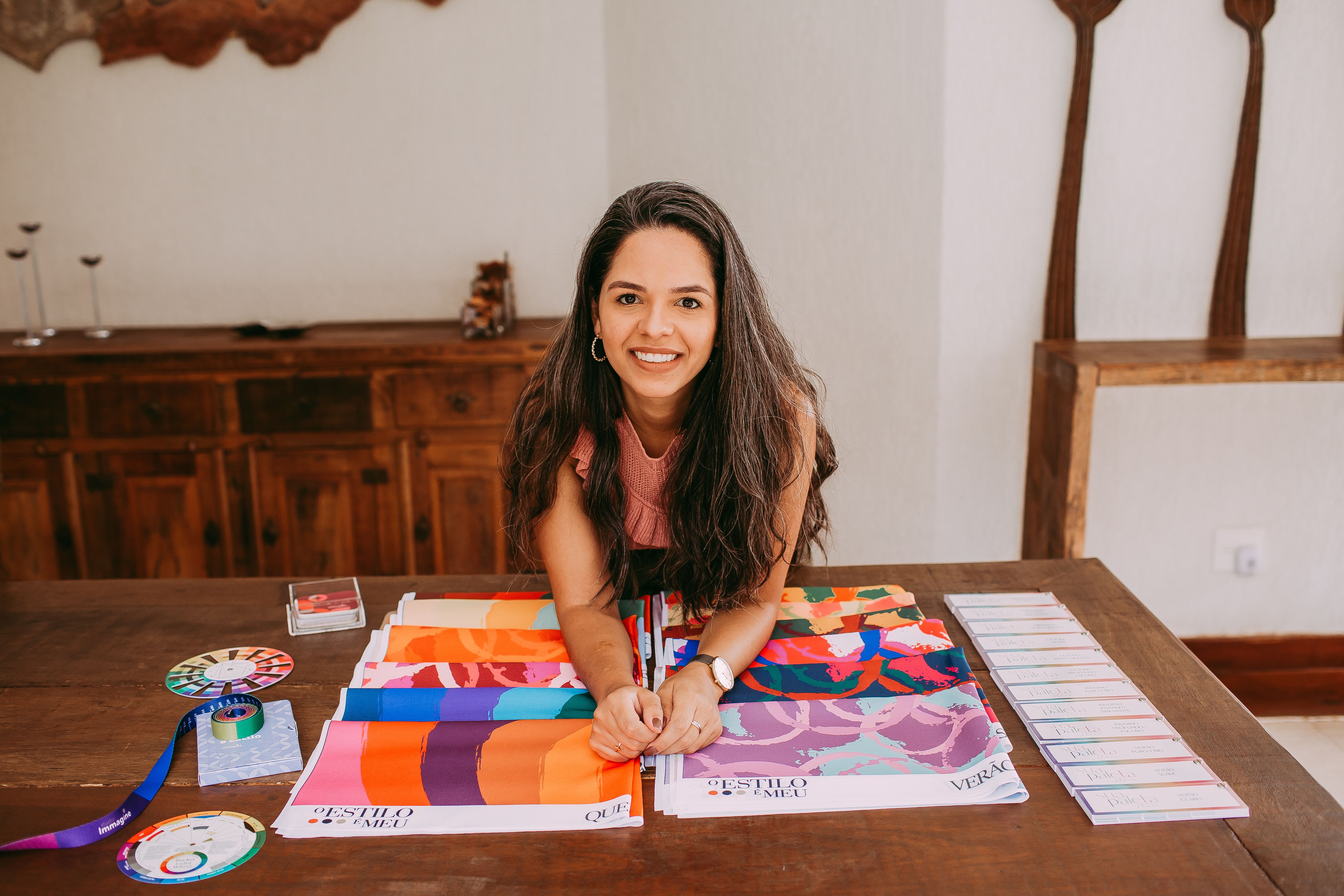 A woman with long hair smiles while posing at a table with colorful fabric samples indoors.