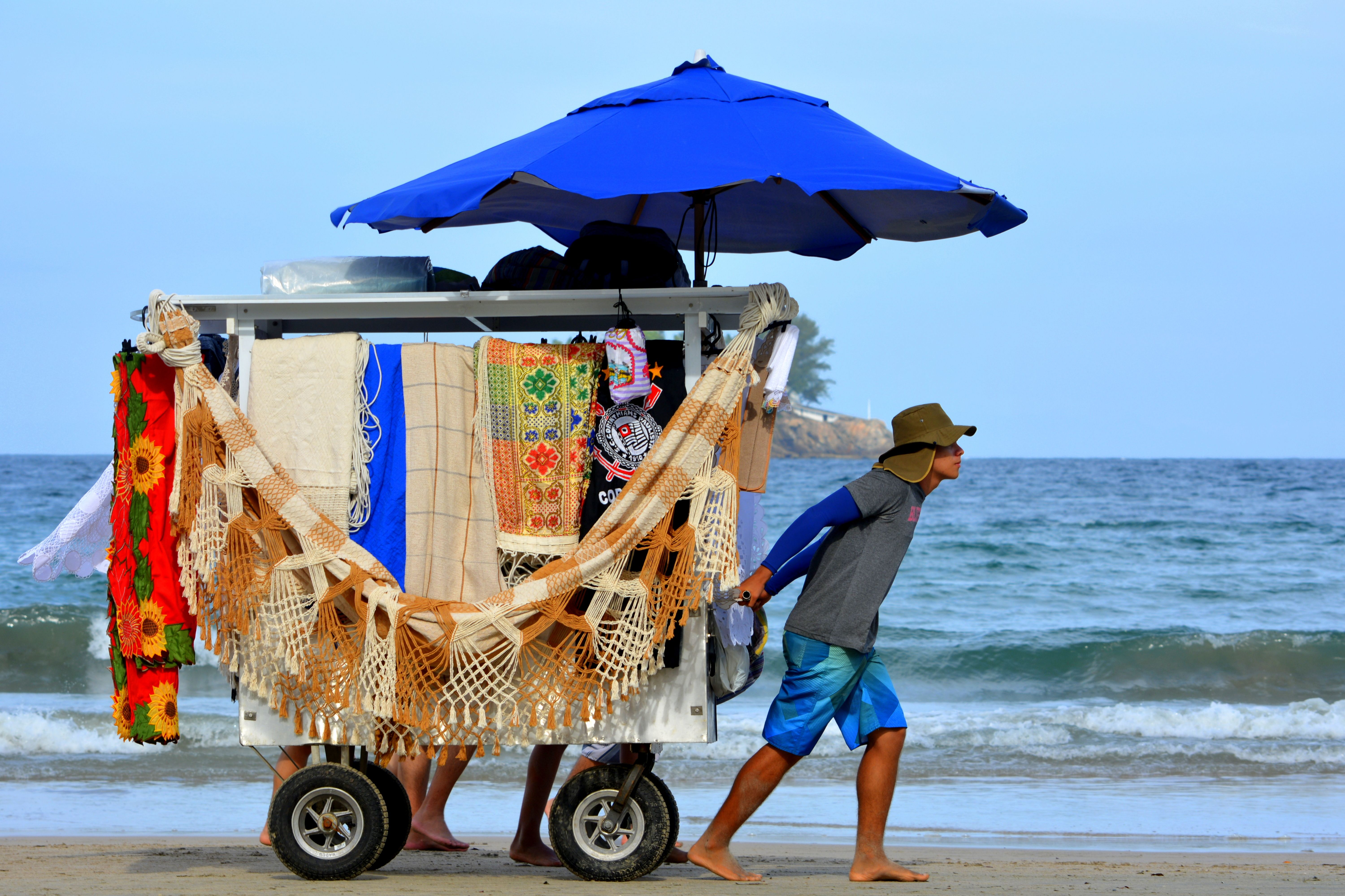 A beach vendor on São Paulo's shore selling colorful textiles from a cart under a blue umbrella.