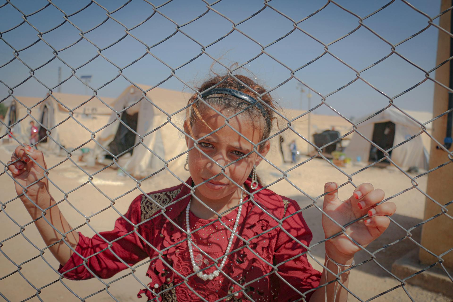 A young girl in a red dress stands behind a chain link fence inside an Idlib refugee camp.