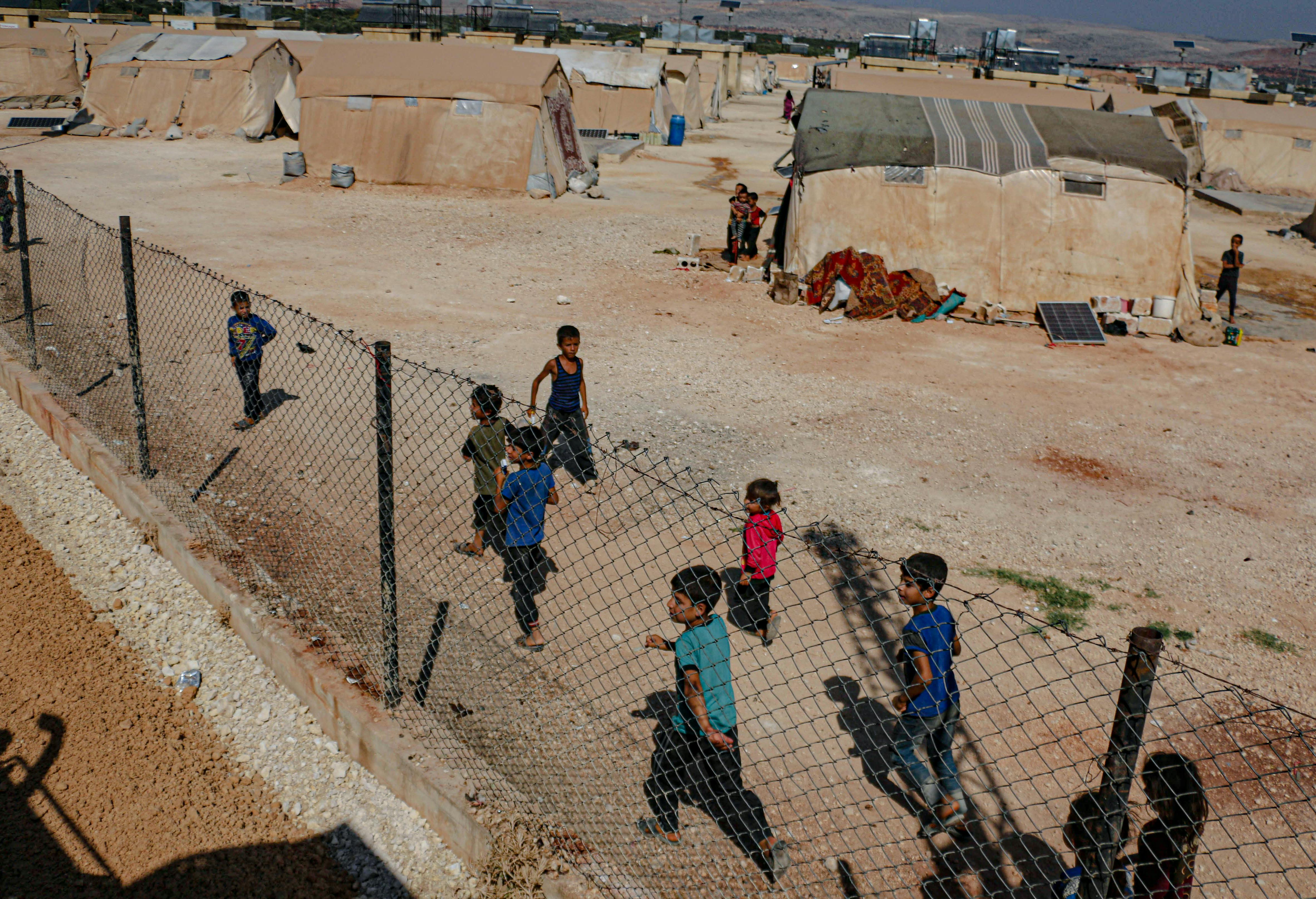 Group of children walk and play near a wire fence in a refugee camp lined with beige tents.