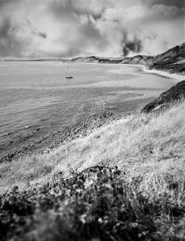 Black and white coastal scene featuring a solitary sailboat under dramatic clouds.