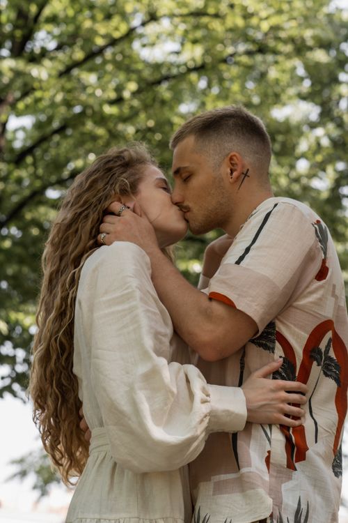 Free A young couple intimately kissing in a sunlit park surrounded by lush greenery. Stock Photo