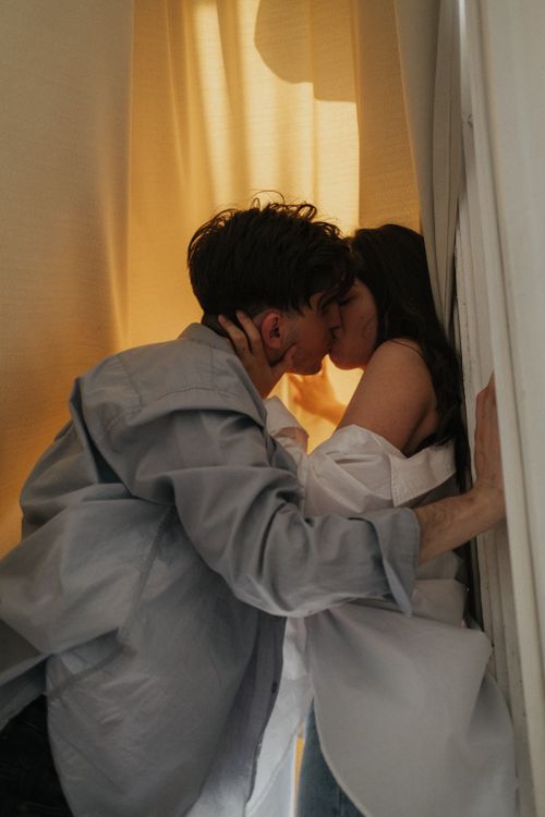 Free Young couple sharing a passionate kiss in a softly lit room with curtains, embodying intimacy and togetherness. Stock Photo