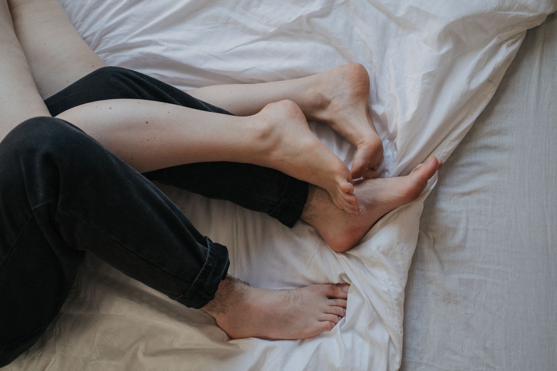 Close-up of a couple's legs and feet intertwined on white bed sheets, conveying intimacy and comfort.