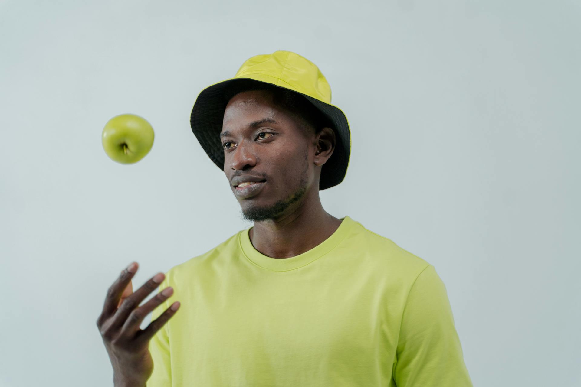 A man wearing a yellow shirt and bucket hat concentrates on balancing a green apple mid-air.