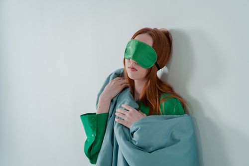 Free A woman with ginger hair rests against a wall, wearing a green sleep mask and holding a blanket. Stock Photo