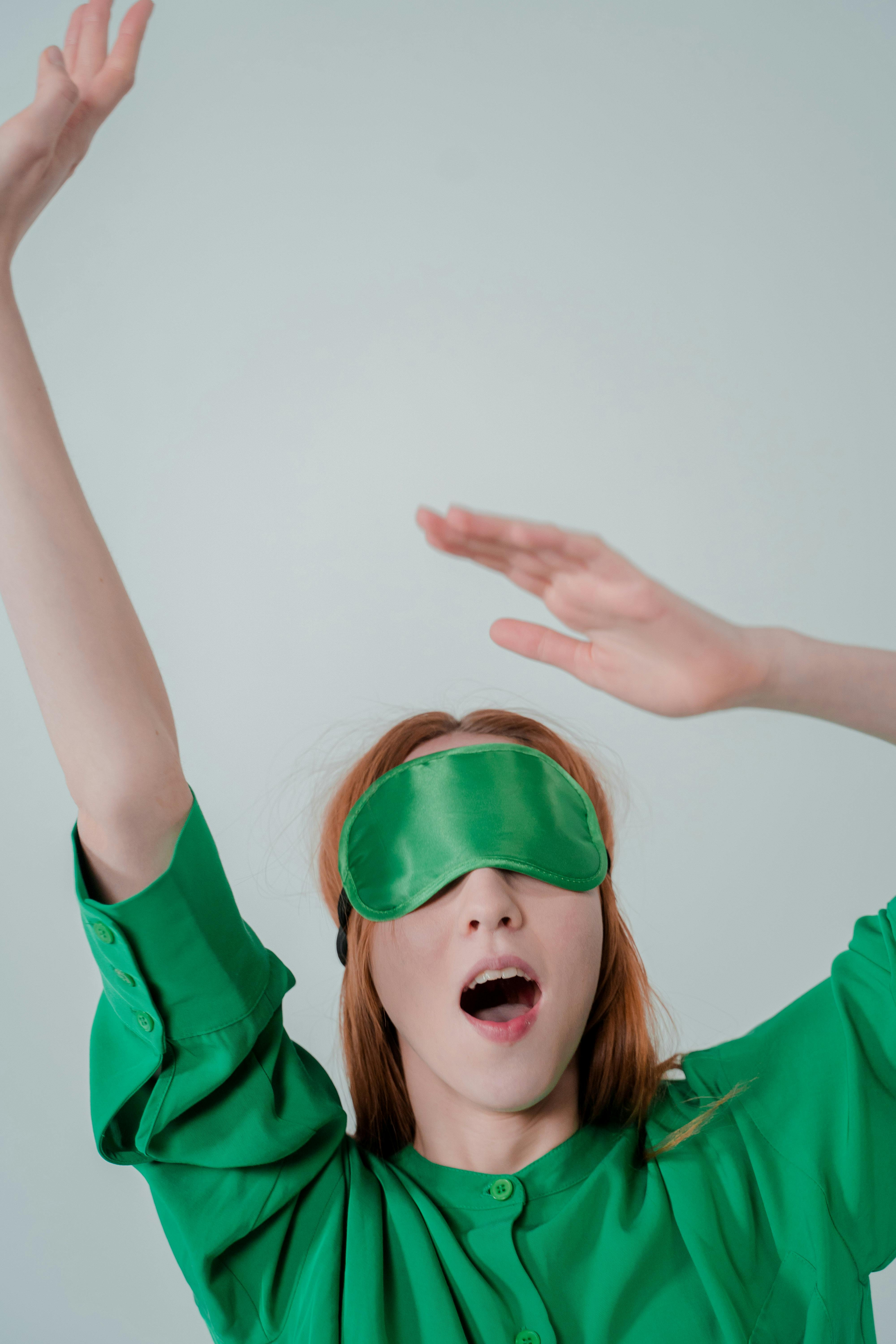 Young woman stretches while wearing a green sleep mask, captured in a studio setting.