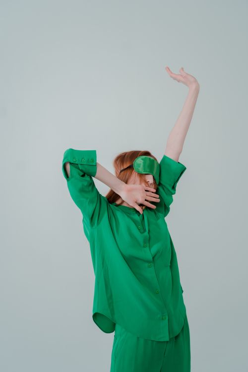 Free Woman in green sleepwear stretching with arms raised, wearing a sleep mask in a minimalist studio setting. Stock Photo