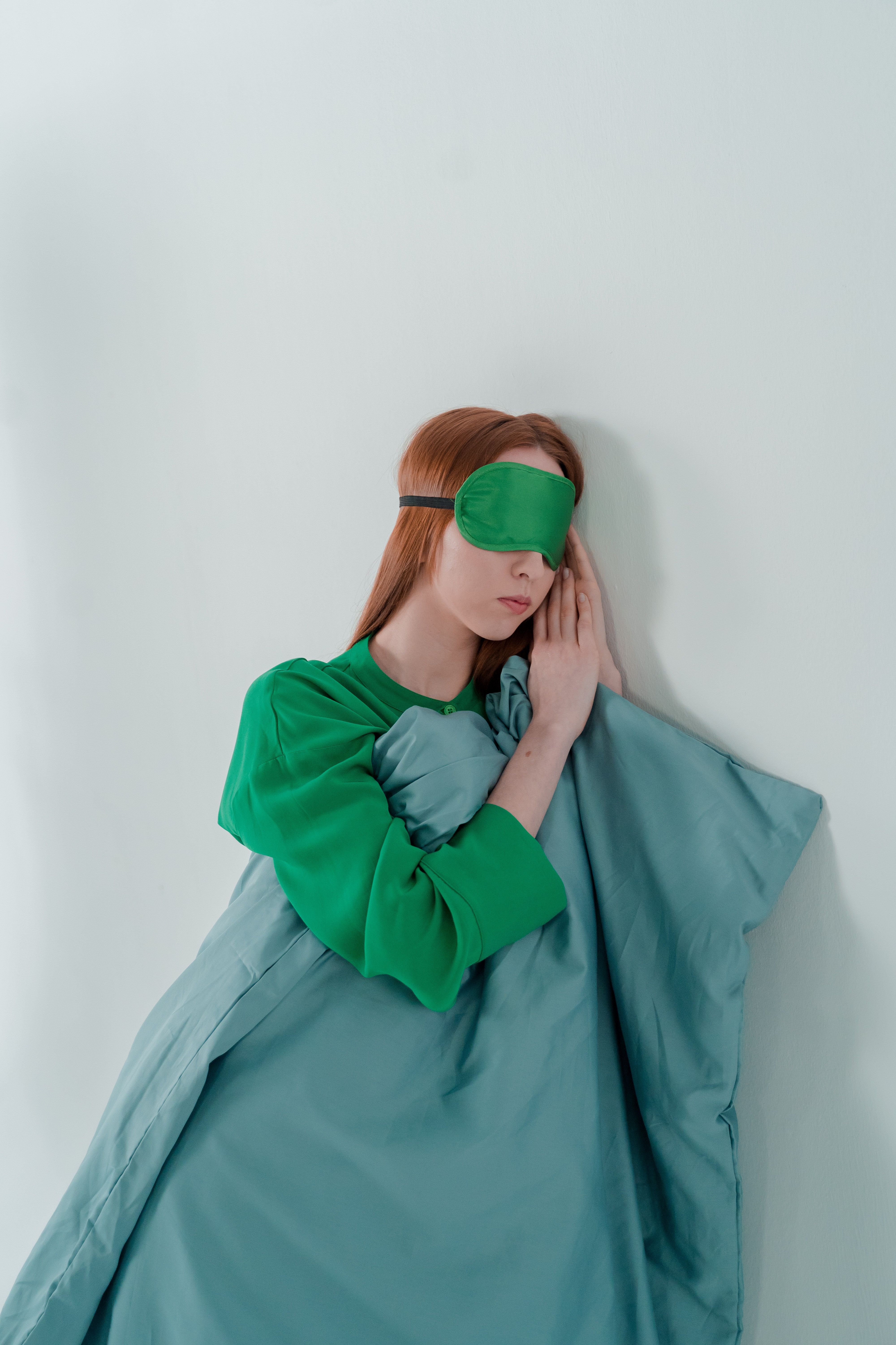 Woman with red hair wearing a green sleep mask and blanket, leaning against a white wall.