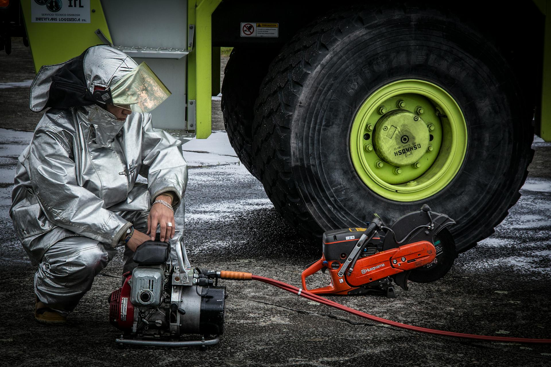 Firefighter in silver protective suit preparing machinery next to a large vehicle tire outdoors.