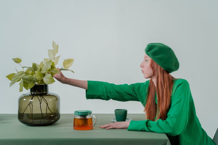 Woman In Green Shirt And Green Beret Sitting By The Table Touching A Plant On Glass Vase