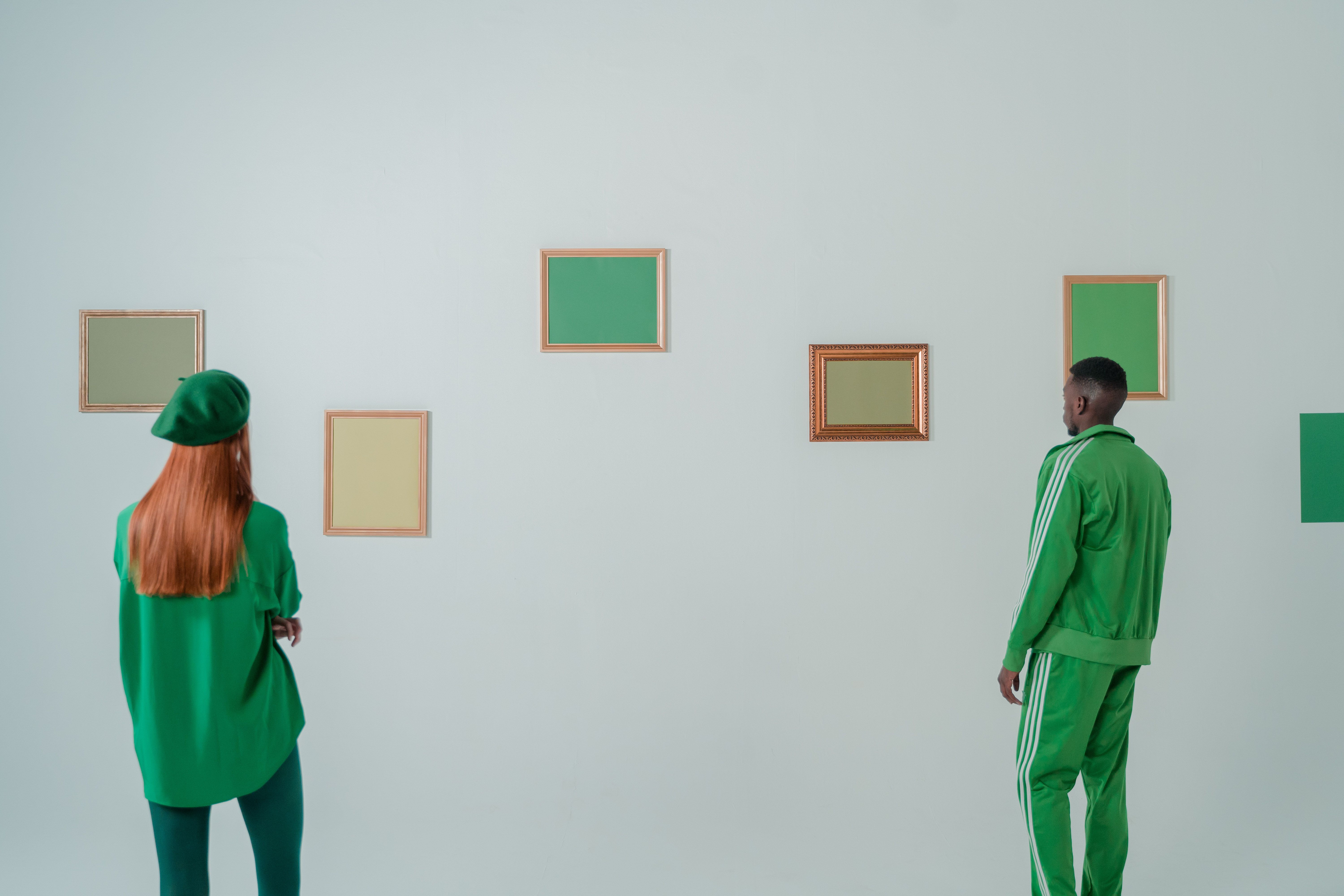 Two individuals in vibrant green outfits examining picture frames on a minimalist wall.