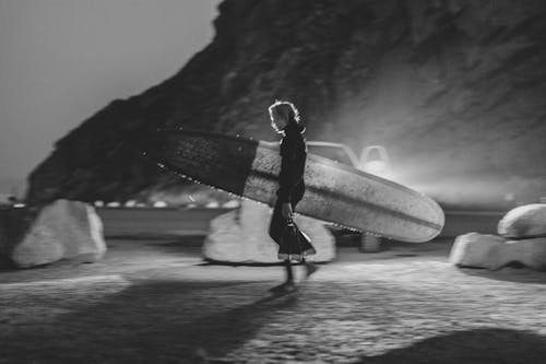 A Grayscale Photo of a Man Carrying a Surfboard