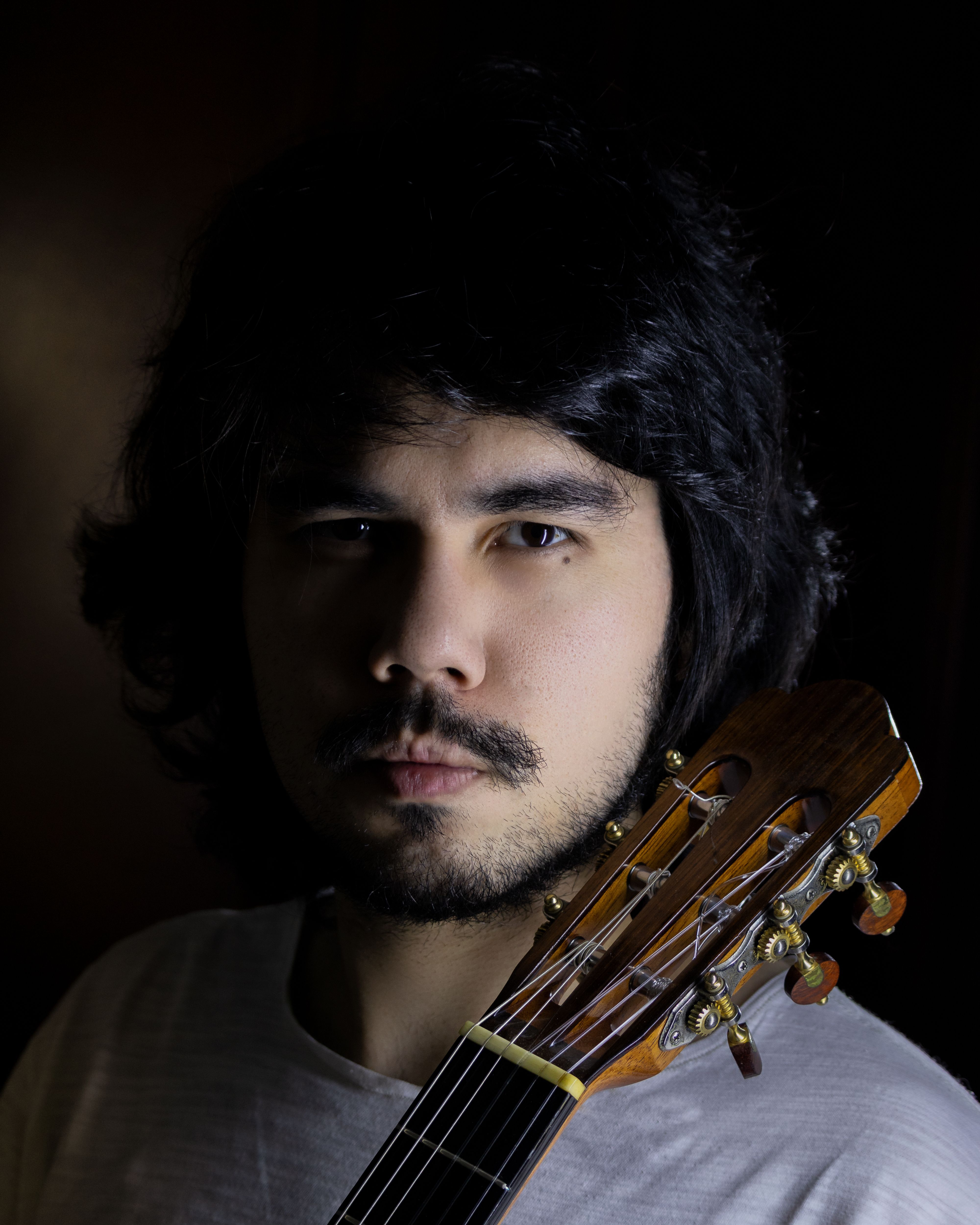 Close-up portrait of a bearded man holding a guitar, captured in dim lighting, showing focus and intensity.