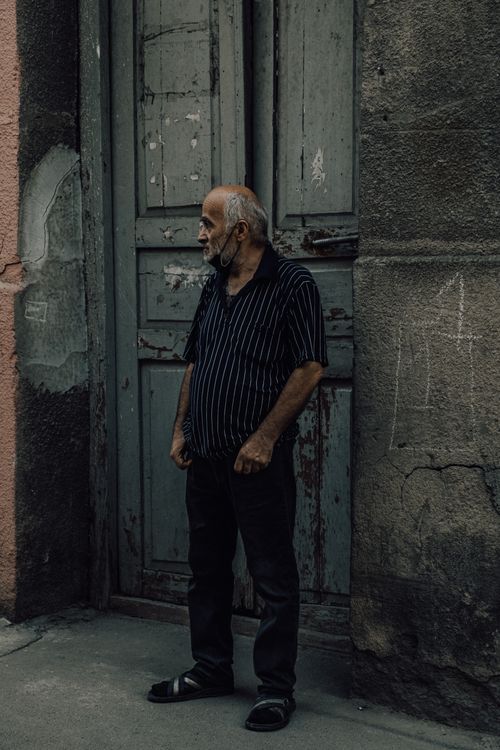Free Elderly man standing by an aged wooden door with a rustic urban backdrop. Stock Photo