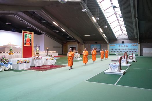 Monks in bright orange robes walk through a decorated hall before a Buddhist ceremony.