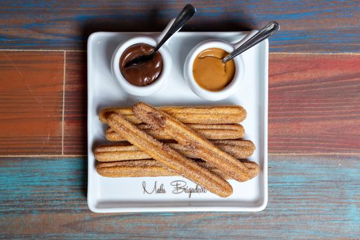 Plate of churros with chocolate and caramel dips on a colorful wooden table.