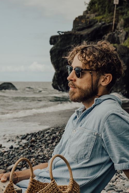 Free Curly-haired man wearing sunglasses relaxing on a pebble beach by the sea. Stock Photo