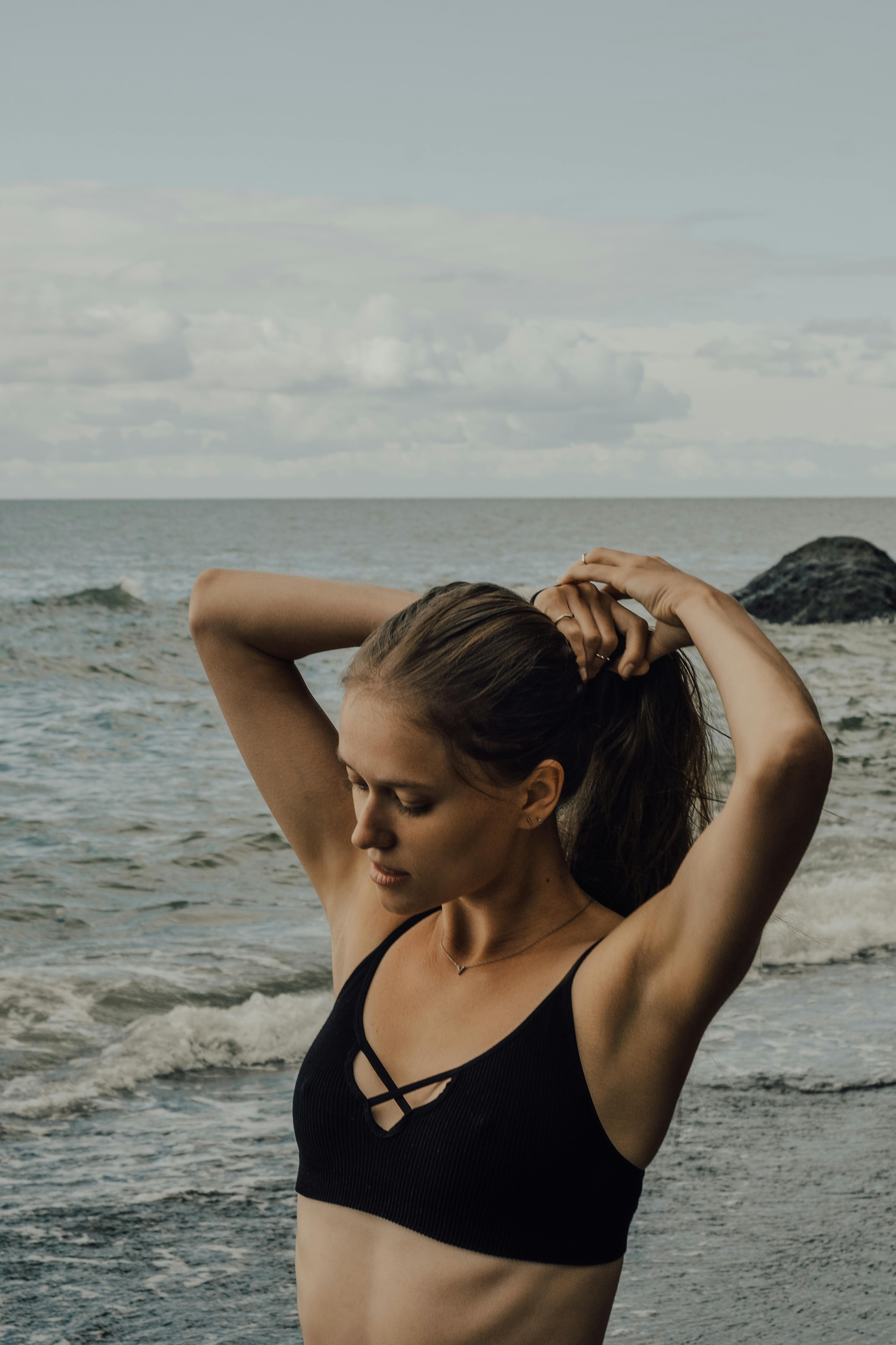 一名穿着黑色比基尼的年轻女子在海滩上扎头发，背景是海浪