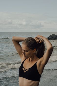A young woman in a black bikini tying her hair on a beach, with waves in the background.