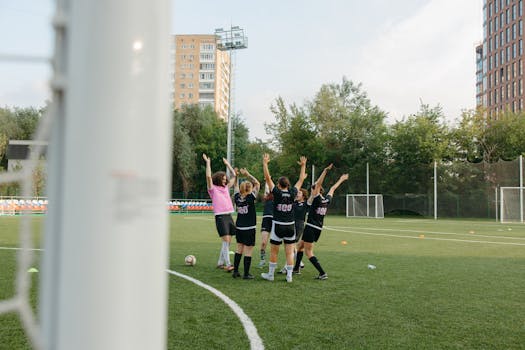 A women's soccer team in black uniforms celebrates a victory on an outdoor soccer field.