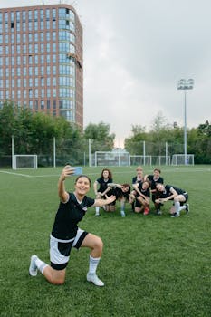 A happy women's soccer team posing for a selfie on a sunny day at an outdoor field.
