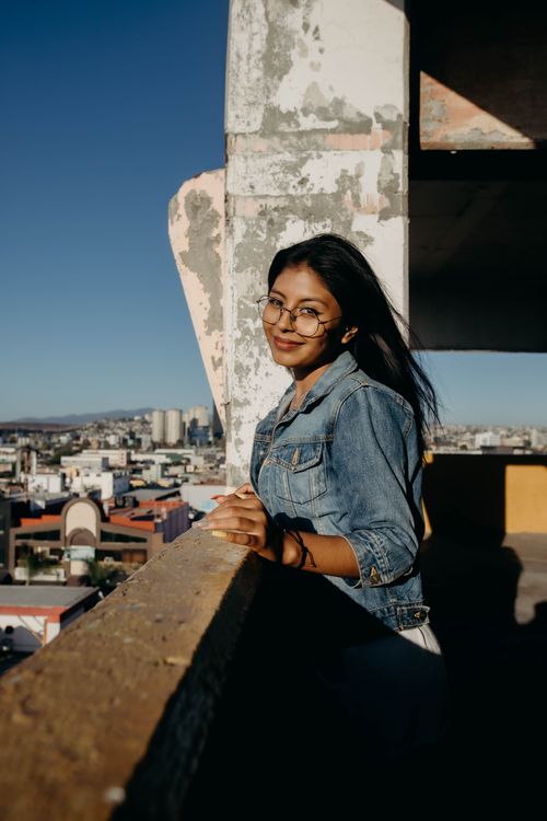 Free Woman leaning on balcony in cityscape, wearing denim jacket and glasses. Stock Photo