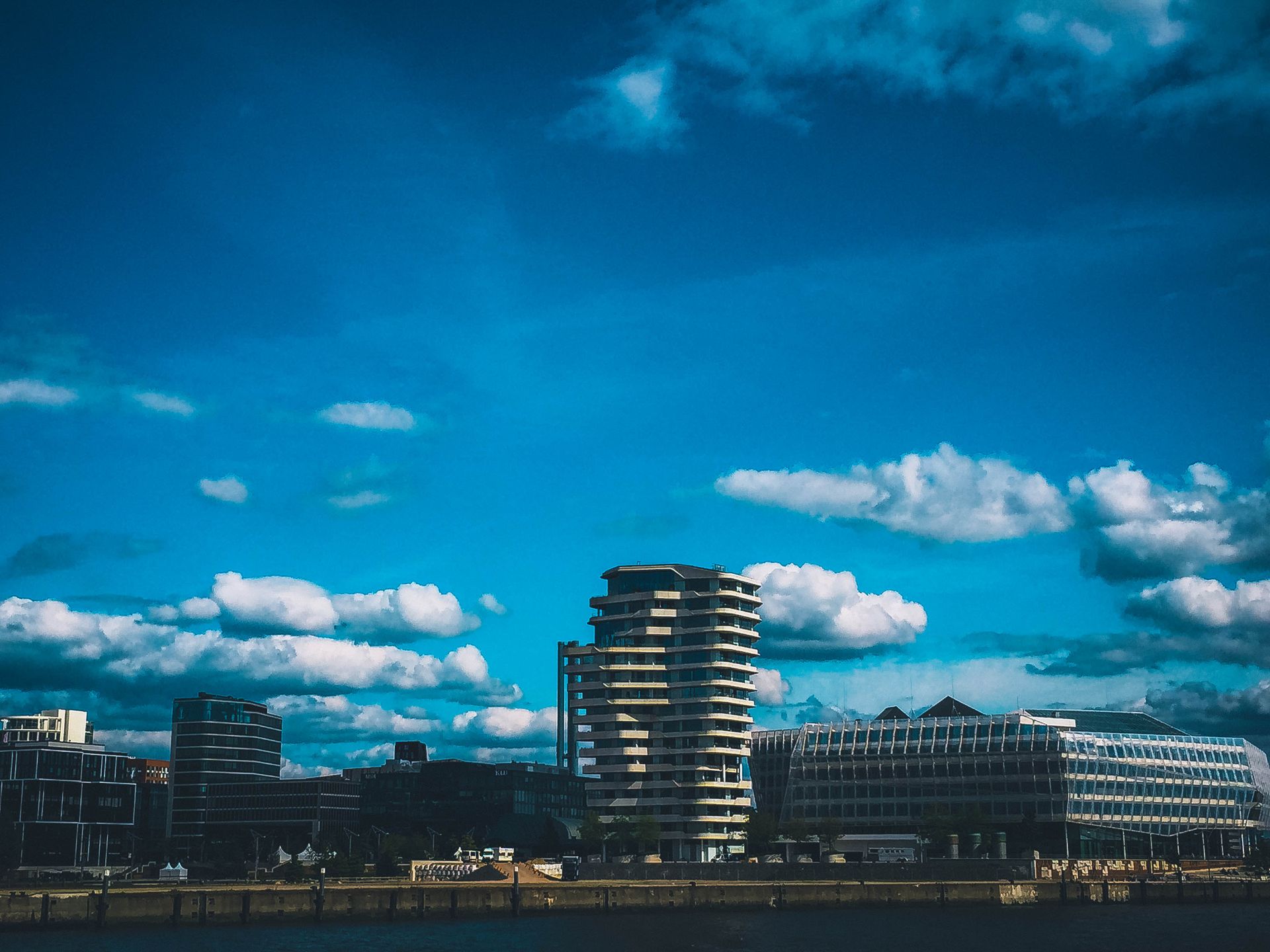 A vibrant city skyline with modern buildings under a clear blue sky, seen from the waterfront.