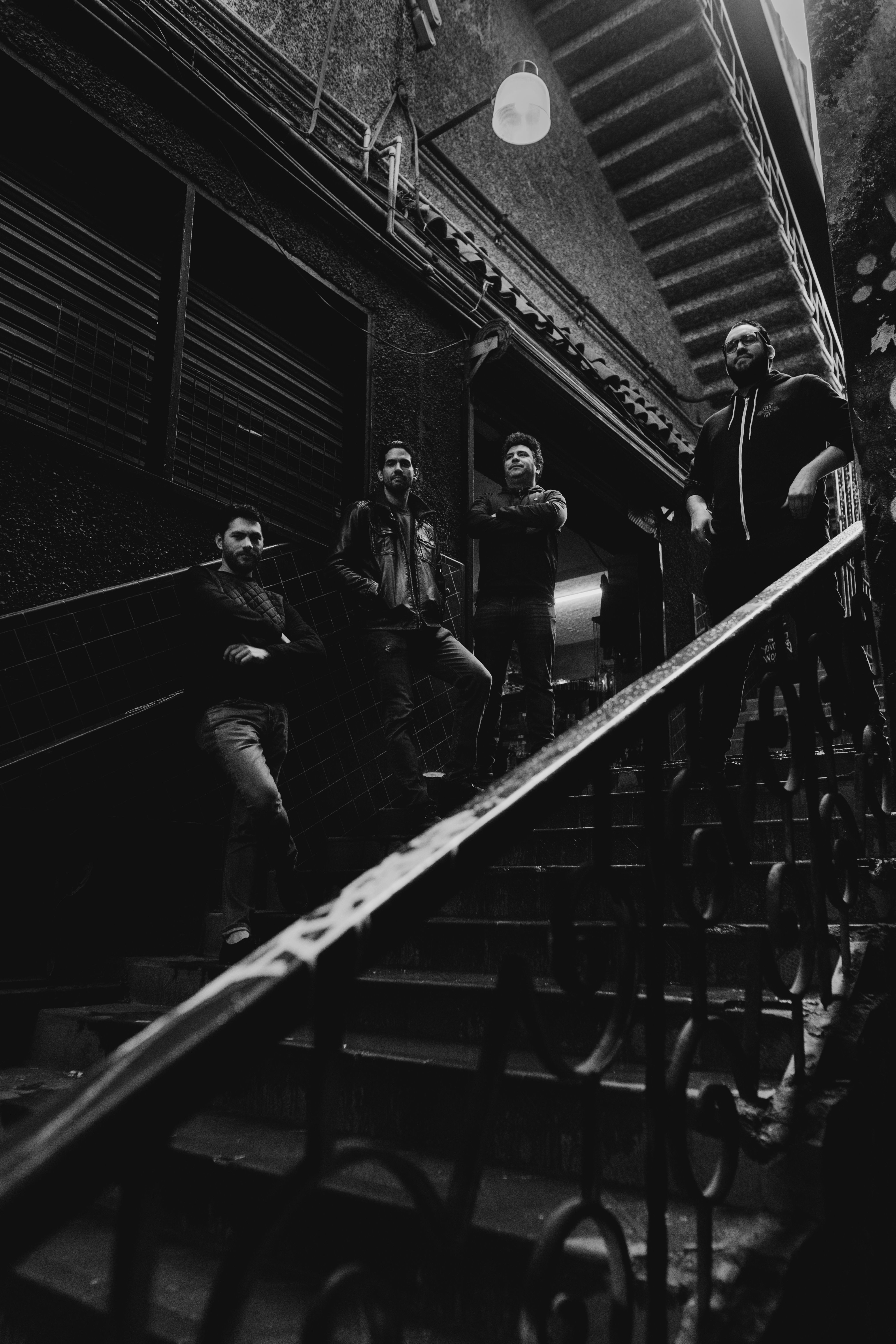 A dramatic black and white photo of four men posing on a dimly lit staircase, adding subcultural flair.